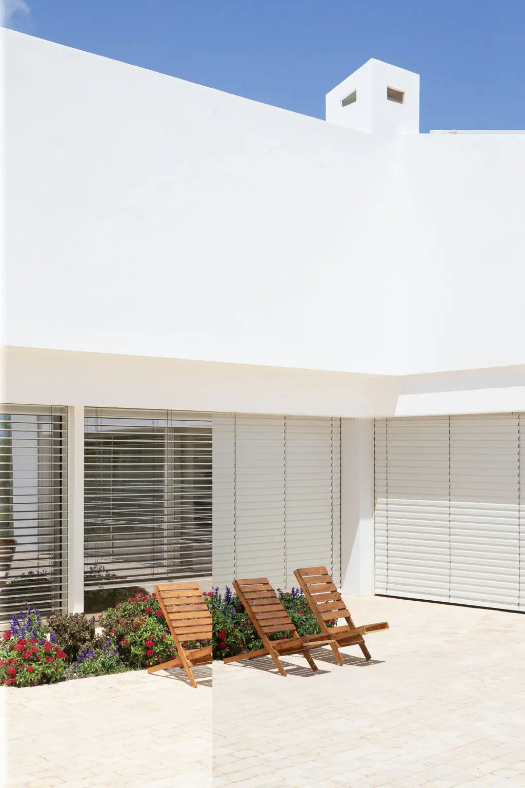 Maison blanche d’allure méditerranéenne sous un ciel bleu, baies du rez-de-chaussée protégées par des brise-soleil orientables à lames blanches ; trois fauteuils en bois sur la terrasse en pierre claire.