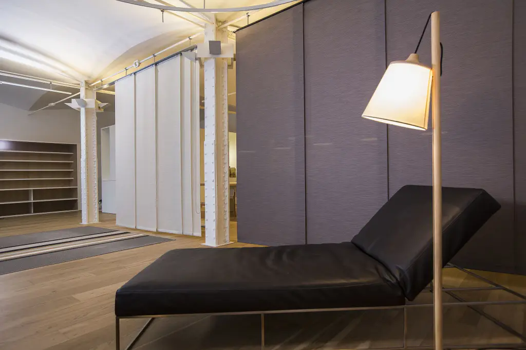 Loft à colonnes métalliques blanches : panneaux japonais blancs et gris anthracite coulissant sur rail au plafond pour cloisonner l’espace ; méridienne en cuir noir et lampadaire en bois au premier plan.