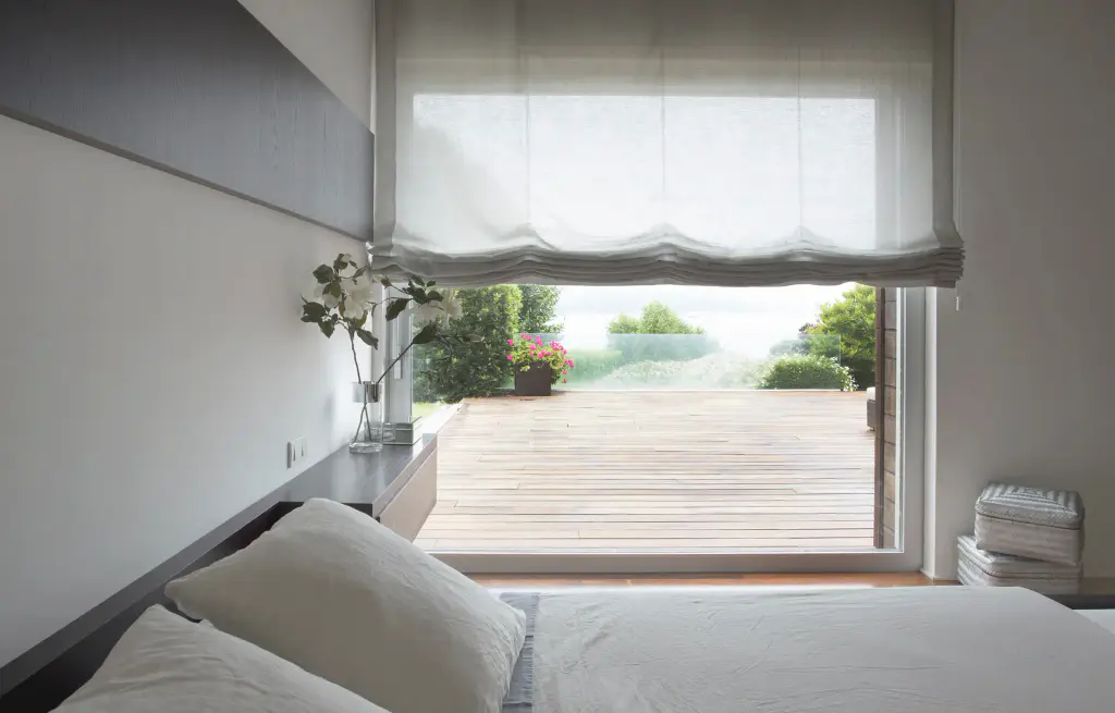 Photographie du fabricant Bandalux : chambre blanche ouverte sur une terrasse en bois et un jardin : un store bateau en voilage blanc, relevé en plis souples en haut de la grande baie ; lit aux draps blancs au premier plan, bouquet dans un vase.
