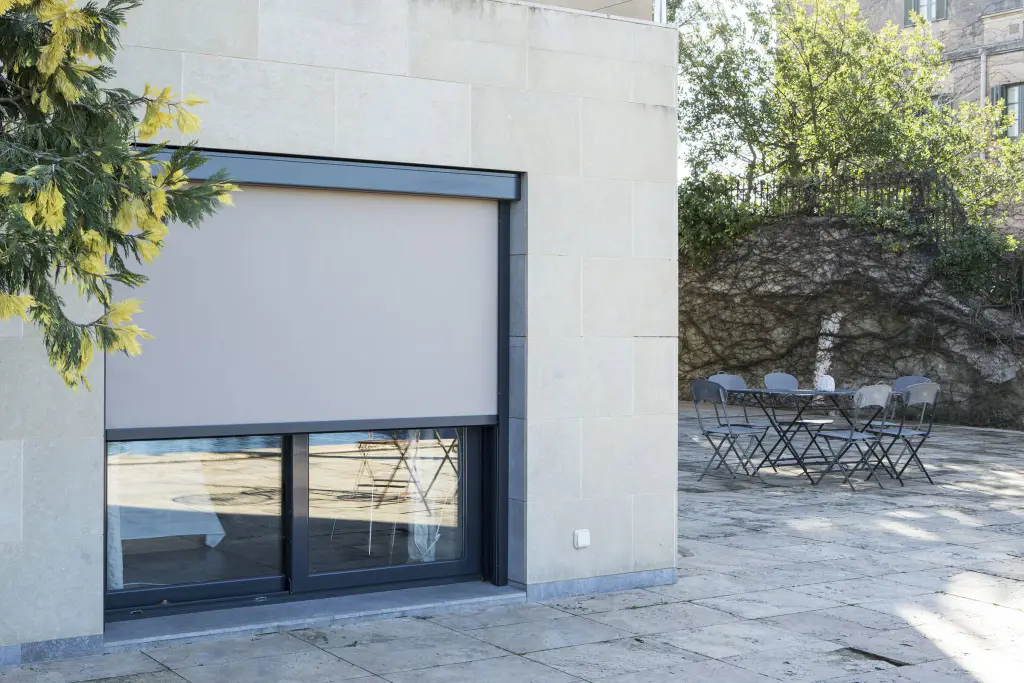 Façade en pierre claire avec un store enrouleur zip beige à coffre et coulisses gris anthracite, descendu aux deux tiers devant une baie coulissante ; terrasse dallée avec table et chaises de jardin à droite, branche de mimosa à gauche.