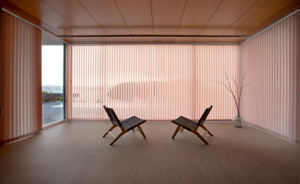 Grande pièce presque vide au plafond de bois : stores à bandes verticales rose pâle fermés sur deux murs vitrés, laissant deviner le paysage ; deux fauteuils bas en bois et cuir tressé se font face au centre.