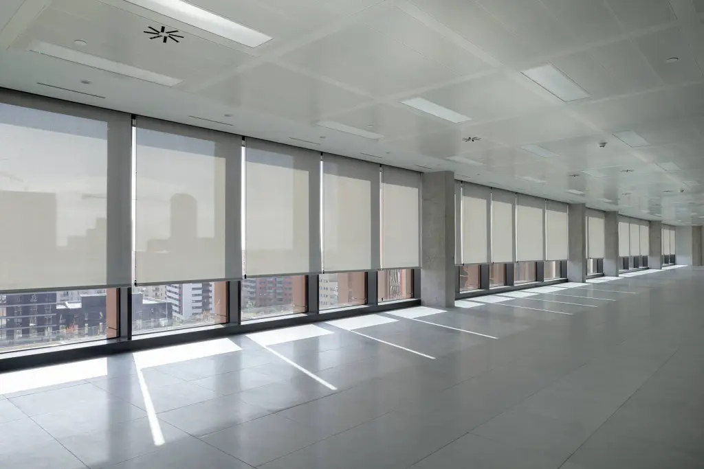 Plateau de bureaux vide, sol de dalles grises : une longue rangée de stores enrouleurs à toile screen claire descendus devant les grandes baies, la ville en transparence ; la lumière dessine des bandes sur le sol.