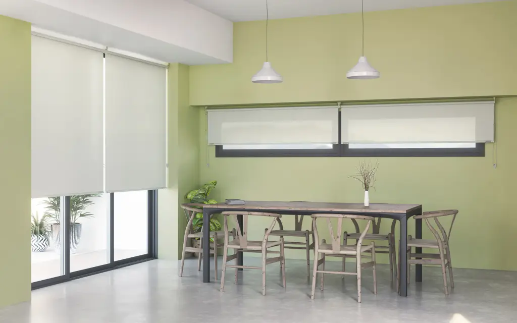 Salle à manger aux murs vert tilleul : deux stores enrouleurs blancs descendus devant une baie vitrée et deux autres relevés au-dessus d’une fenêtre en bandeau ; longue table et chaises en bois clair, deux suspensions blanches.