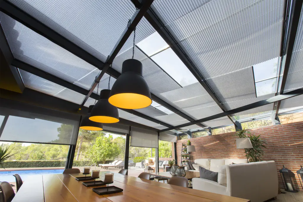Véranda à structure noire vue en contre-plongée : stores plissés gris clair tendus sous chaque travée de la toiture vitrée, stores enrouleurs sur les côtés ; trois suspensions noires au-dessus d’une table en bois, piscine et pins au-dehors.