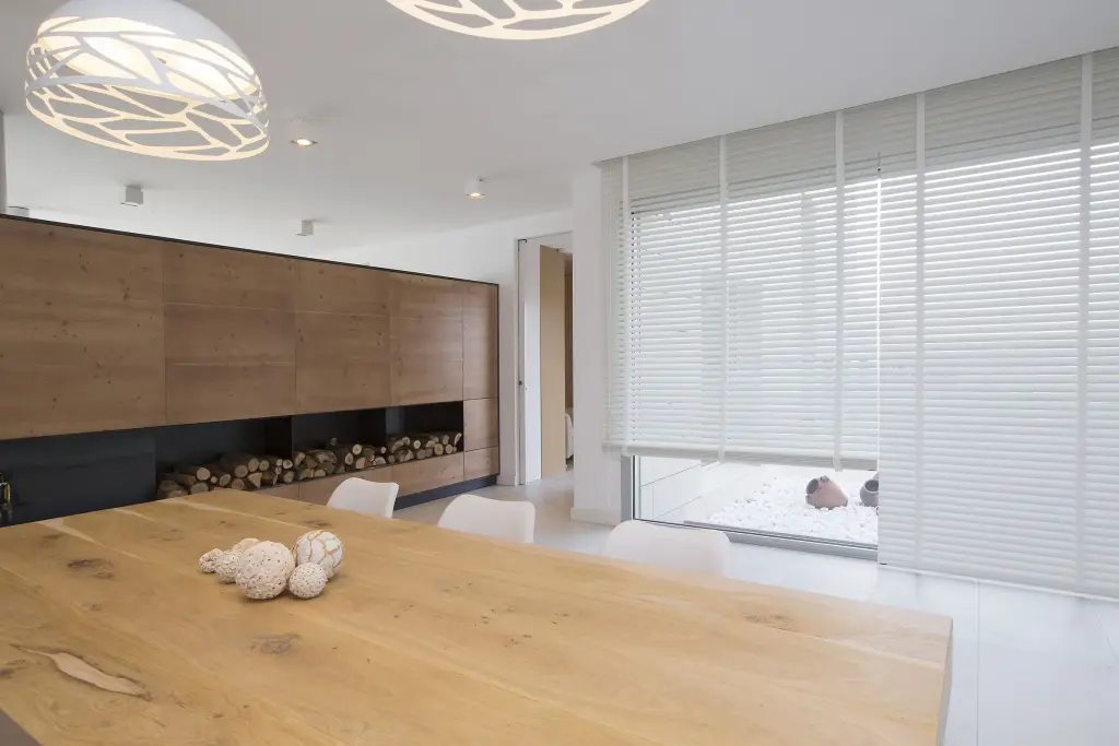 Salle à manger blanche : stores vénitiens à lames blanches sur toute la hauteur de la baie d’angle, grande table en chêne massif au premier plan, mur de rangements en bois avec niche à bûches, suspensions ajourées.