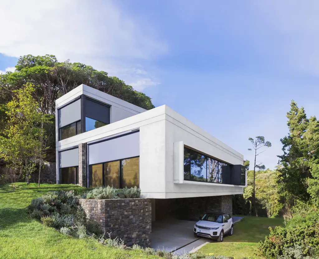 Villa cubique en béton blanc sur un terrain en pente boisé : stores enrouleurs extérieurs gris descendus à mi-hauteur sur les baies des deux niveaux ; soubassement en pierre sèche, un 4x4 blanc garé sous le porte-à-faux.