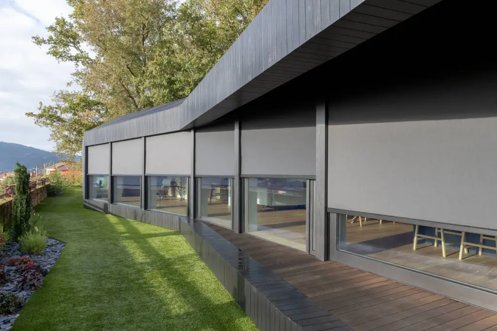 Maison contemporaine de plain-pied au bardage noir, vue en enfilade depuis le jardin : six stores enrouleurs zip gris clair descendus aux trois quarts devant les baies vitrées, terrasse en bois et pelouse au premier plan.
