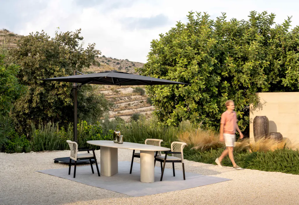 Jardin sec en restanques, sol de gravier clair : un parasol déporté à toile noire au-dessus d’une table ovale et de quatre chaises ; un homme passe devant un mur ocre et de grandes jarres.