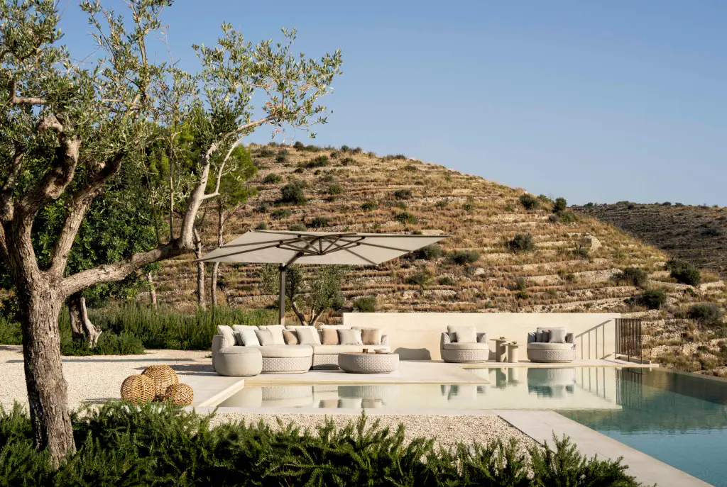 Piscine à débordement au pied d’une colline en restanques : un parasol déporté à toile claire au-dessus d’un salon tressé blanc, un olivier au premier plan.