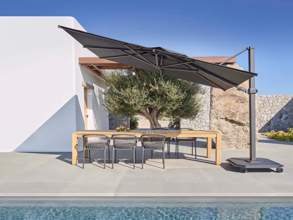 Terrasse d’une maison blanche au bord d’une piscine : un grand parasol déporté à toile anthracite, incliné, abrite une longue table en teck et ses chaises tressées ; derrière, un olivier, un rocher et des murets de pierre sèche.