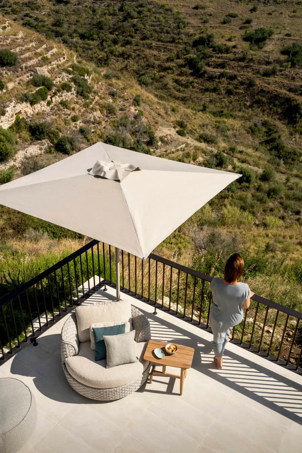 Terrasse en étage au-dessus d’une colline en restanques : un parasol carré à toile blanche ombre un fauteuil rond et une table basse ; une femme s’accoude au garde-corps.