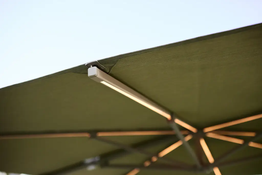 Vue de dessous d’un parasol à toile vert olive : les baleines éclairées par des rubans lumineux, un petit boîtier au bout du bras.