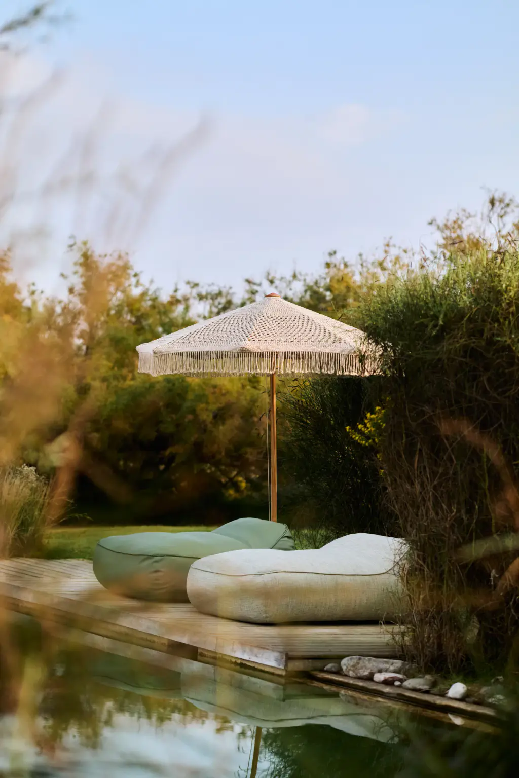 Au bord d’un bassin naturel bordé de roseaux : un parasol rond en bois à franges de macramé écru, au-dessus de deux grands poufs posés sur un ponton.