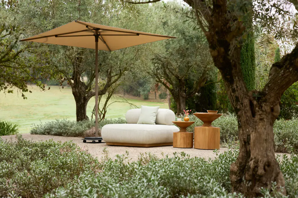 Clairière entre de vieux oliviers : un parasol carré à mât central et toile sable abrite une banquette blanche et deux tables basses en bois tourné, au milieu d’un massif bas.
