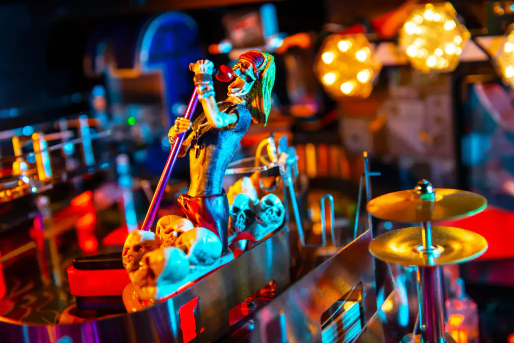 Sur le plateau du flipper Guns N’ Roses, une batterie miniature avec ses cymbales dorées, devant une sculpture de squelettes et des lumières rondes.
