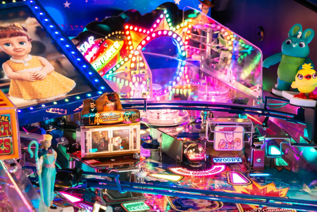 Le fond du plateau du flipper Toy Story 4 : un décor de fête foraine éclairé de rose et de bleu, avec des figurines de jouets de part et d’autre.