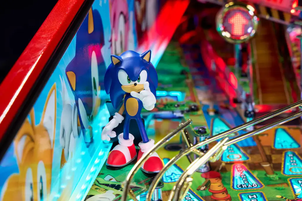 Sur le plateau du flipper Sonic, la figurine bleue du personnage, debout près d’une rampe en fil d’acier, dans la lumière des inserts.
