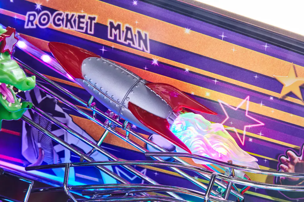 Sur le plateau du flipper Elton John, une fusée chromée sculptée, ses flammes en LED, sous un panneau « Rocket Man ».