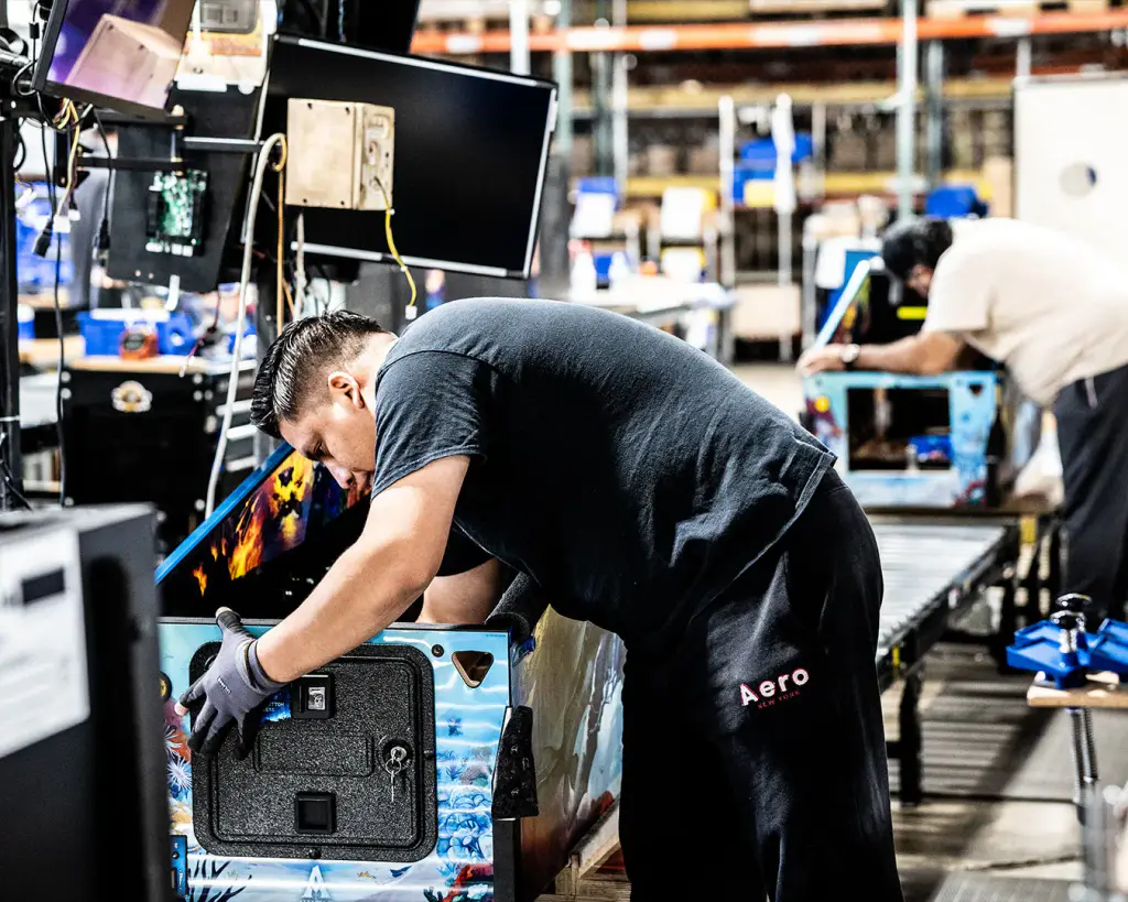 Dans l’usine Jersey Jack Pinball, un ouvrier ganté, penché, pose la porte de monnayeur noire sur la face avant d’une caisse de flipper ; convoyeur à rouleaux et rayonnages au fond.