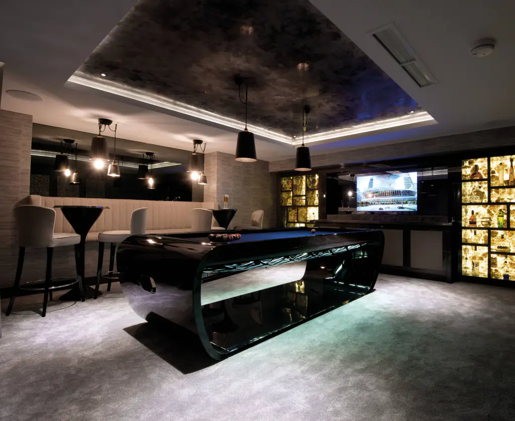 Billard noir laqué aux flancs arrondis, éclairé par-dessous, au centre d’un salon-bar sombre : moquette grise, banquette claire, suspensions noires et vitrine à bouteilles rétroéclairée.
