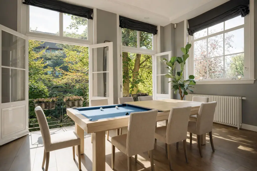 Billard en bois clair à drap bleu, à moitié recouvert de son plateau de table en bois, entouré de six chaises beiges, dans une salle à manger grise à grandes fenêtres ouvertes sur des arbres.