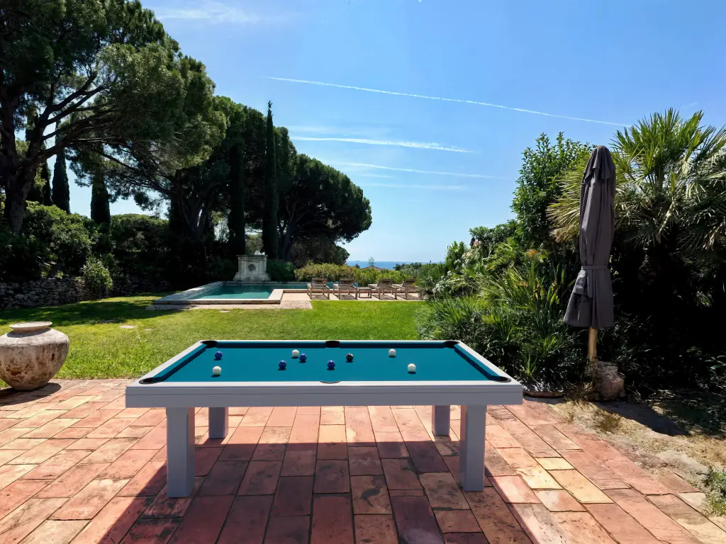 Billard d’extérieur blanc à drap bleu pétrole, billes bleues et blanches, posé sur une terrasse en tomettes devant une pelouse, une piscine, des pins et des cyprès, la mer à l’horizon ; vue de face, parasol fermé à droite.