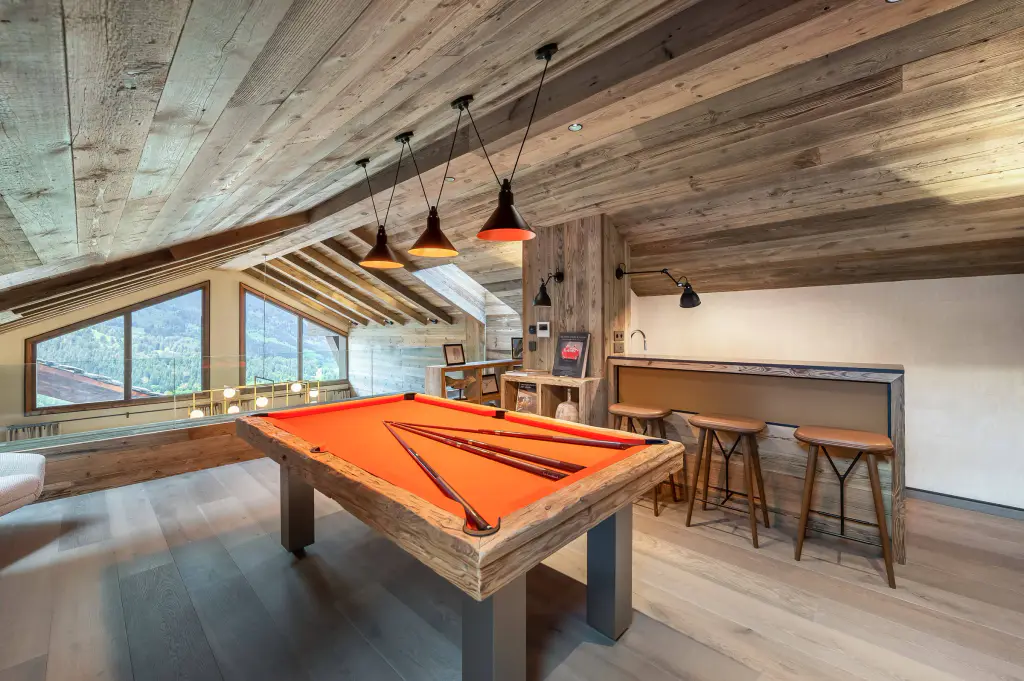Billard au cadre en vieux bois, pieds métal gris, drap orange, quatre queues posées dessus, sous les combles lambrissés d’un chalet avec trois suspensions noir et orange, un petit bar et trois tabourets ; baie sur la montagne à gauche.