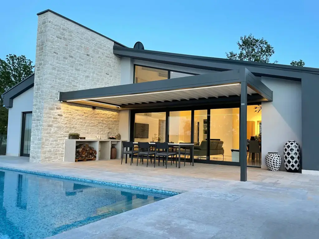 Pergola à toile gris anthracite adossée à une villa contemporaine, à un seul montant, toile blanche et bandeau LED allumé à la tombée du jour ; table et chaises noires devant une grande baie vitrée éclairée, mur de pierre claire, piscine au premier plan.