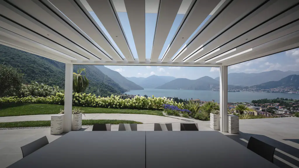 Vue depuis le dessous d’une pergola bioclimatique blanche dont les lames sont entrouvertes sur le ciel ; table grise au premier plan, massifs d’hortensias, vue plongeante sur un lac entouré de montagnes.