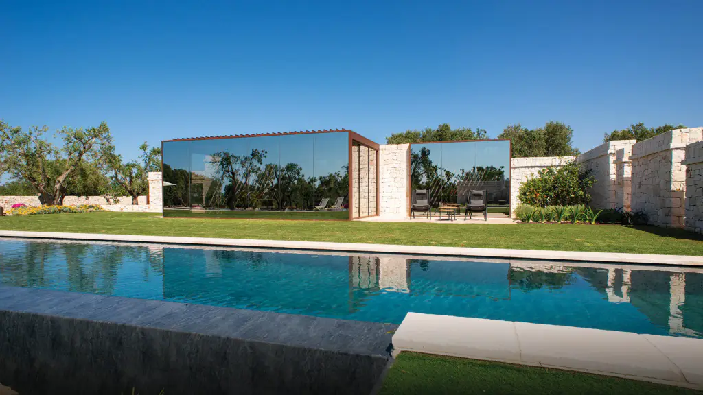 Pergola bioclimatique à vitrages miroir et cadre couleur corten, posée entre des murs de pierre sèche : les vitres reflètent les oliviers. Piscine au premier plan, pelouse, deux fauteuils sous l’auvent voisin, ciel bleu sans nuage.