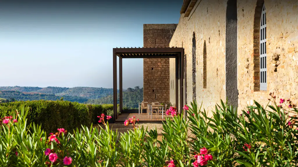 Pergola bioclimatique brun foncé adossée à un long bâtiment ancien en pierre et brique, abritant une table et des chaises sur une terrasse en bois ; lauriers-roses en fleurs au premier plan, collines au loin.
