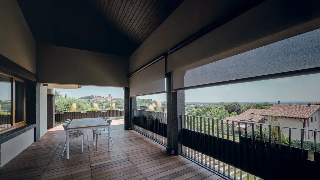 Terrasse couverte d’une villa, plancher en bois et plafond lambrissé sombre : deux stores verticaux à toile gris foncé ajourée, à demi descendus dans les ouvertures latérales, laissent voir les collines et un village ; table blanche et chaises.