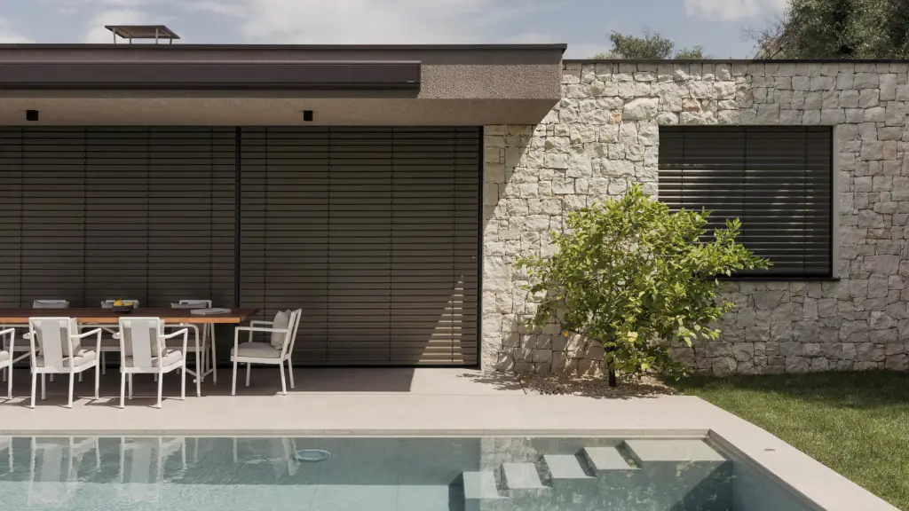 Maison en pierre claire vue de face côté piscine : la grande baie de la terrasse couverte et la fenêtre du mur de pierre sont fermées par des brise-soleil orientables à lames brun foncé ; table et chaises blanches, citronnier, bassin au premier plan.