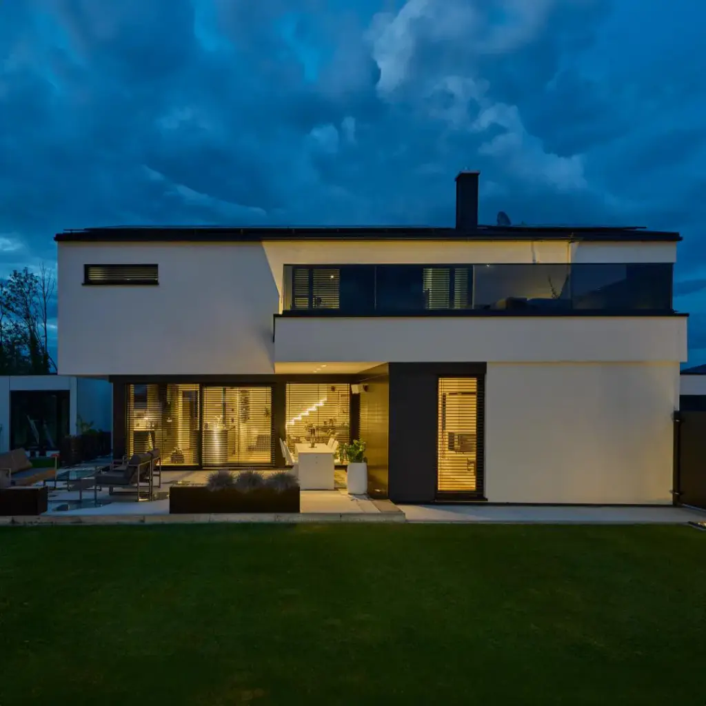 Maison blanche à toit plat à la tombée de la nuit : l’intérieur éclairé se voit à travers des brise-soleil orientables à lames horizontales entrouvertes, au rez-de-chaussée et à l’étage ; pelouse au premier plan, ciel bleu nuit.