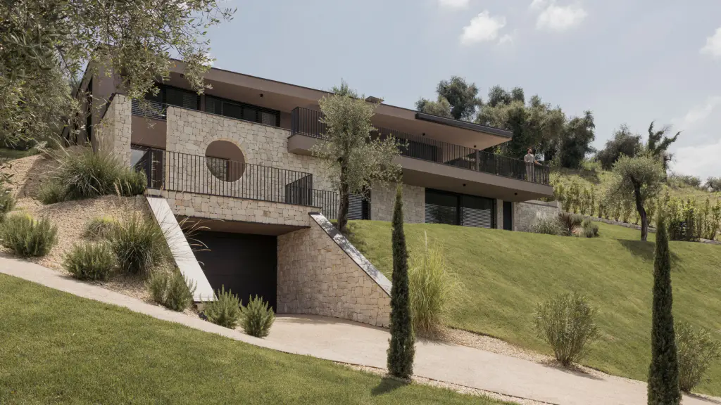 Maison contemporaine en pierre claire à flanc de colline, deux niveaux à toits plats débordants : les baies sont équipées de brise-soleil orientables sombres ; oliviers, cyprès et pelouse en pente.