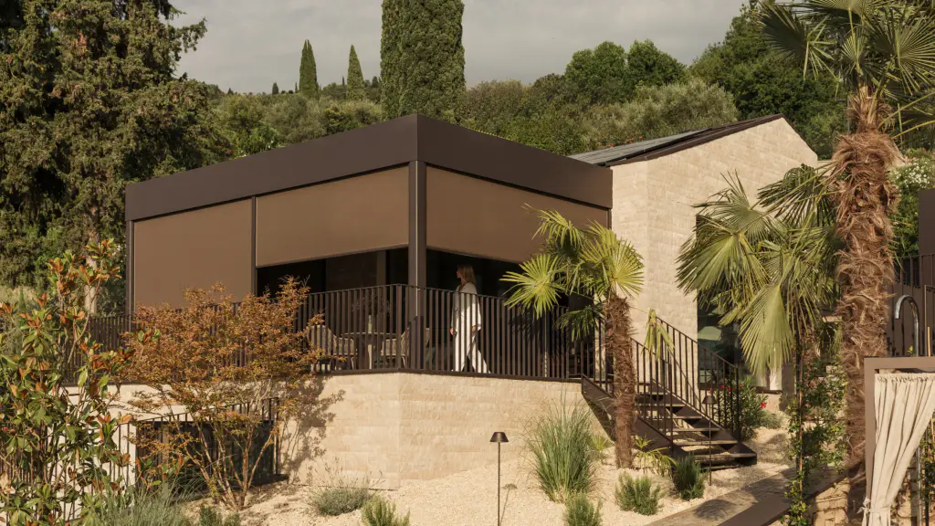 Terrasse couverte d’une maison en pierre, fermée sur deux côtés par des stores toiles verticaux brun foncé à demi descendus dans une structure sombre ; garde-corps à barreaux, palmiers et cyprès, une silhouette au loin sur la terrasse.