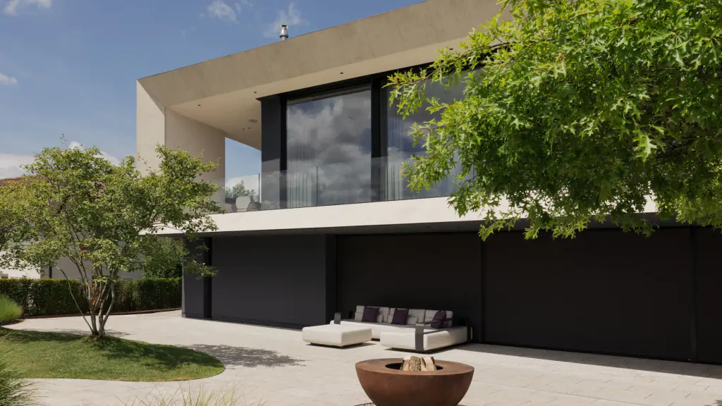 Villa contemporaine en béton clair : au rez-de-chaussée, une longue façade entièrement fermée par des stores toiles noirs ; à l’étage, une grande baie vitrée d’angle ; terrasse dallée avec canapé blanc et brasero en acier corten, érables.