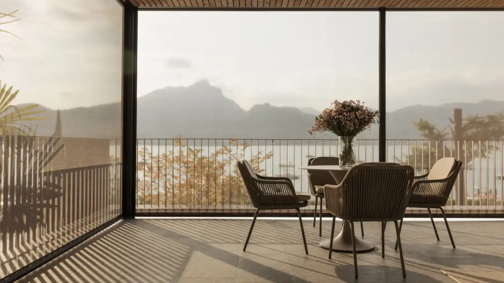 Vue de l’intérieur d’une terrasse couverte : stores toiles verticaux micro-perforés descendus, laissant voir le lac et les montagnes à contre-jour, table ronde avec bouquet et quatre fauteuils tressés, ombres rayées au sol.