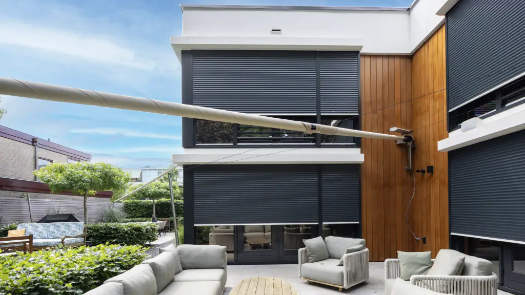 Maison contemporaine blanche à bardage bois vertical : les grandes baies des deux niveaux sont fermées aux trois quarts par des volets roulants gris anthracite ; terrasse avec salon de jardin clair, voile d’ombrage enroulée au premier plan.