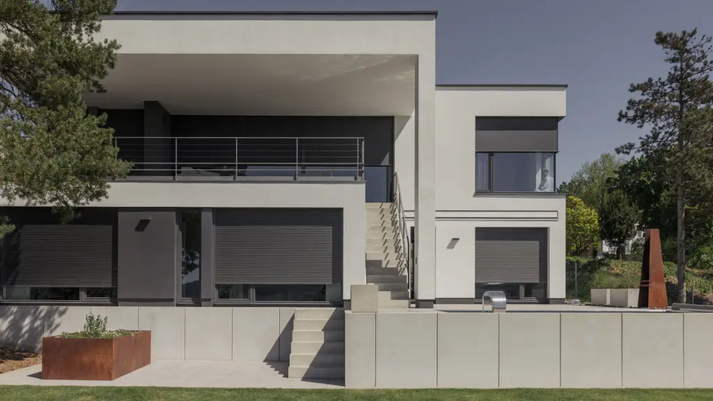 Maison cubique en enduit gris clair vue de face : volets roulants gris anthracite à demi baissés sur les baies du rez-de-chaussée, stores toiles à l’étage, escalier extérieur en béton, jardinière en acier corten, pins.