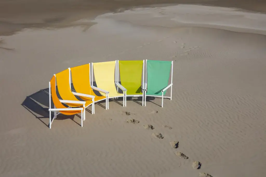 Cinq chaises à structure tubulaire blanche et assise en toile tendue, du orange au vert en passant par le jaune, disposées en arc sur une plage de sable ; traces de pas au premier plan.
