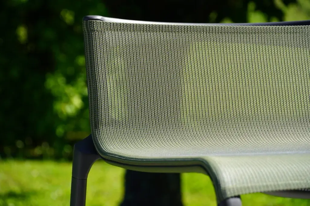 Gros plan sur le dossier d’un fauteuil d’extérieur : toile tendue ajourée vert sauge sur un cadre gris anthracite, pelouse et feuillage flous à l’arrière-plan. Détail de matière.