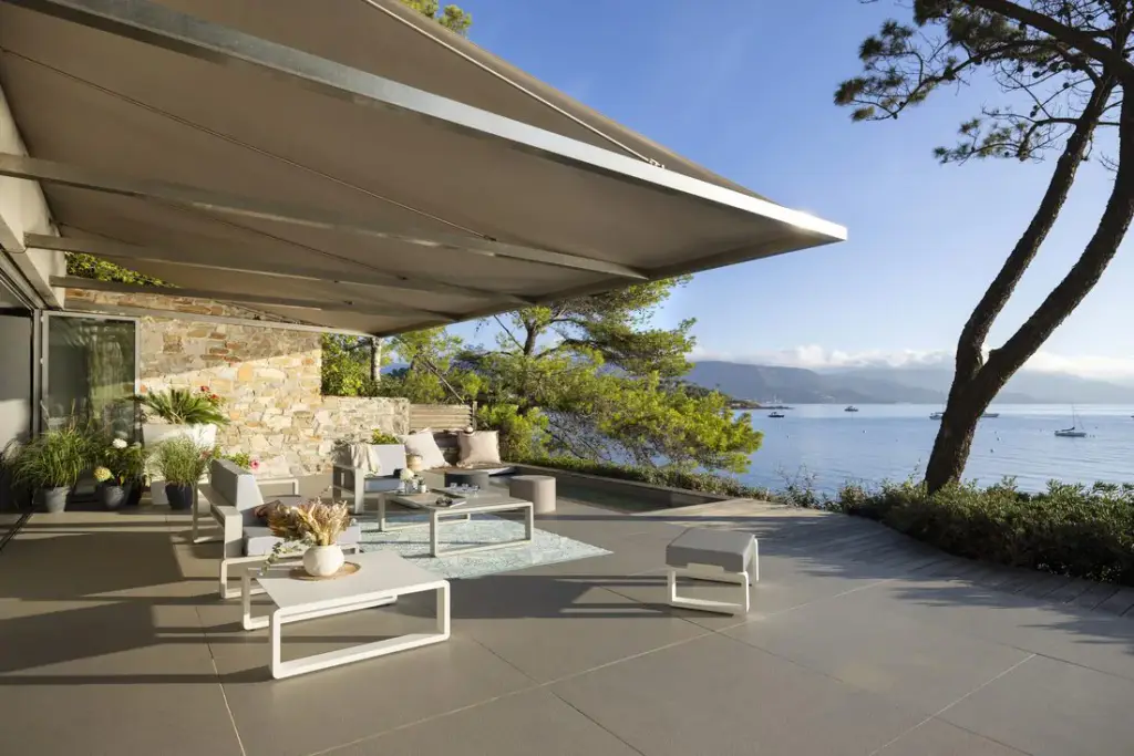 Grand store banne à toile grège déployé sur une terrasse dallée en bord de mer ; salon bas blanc, tapis bleu, mur en pierre, pins parasols, baie avec voiliers au mouillage et collines au loin.