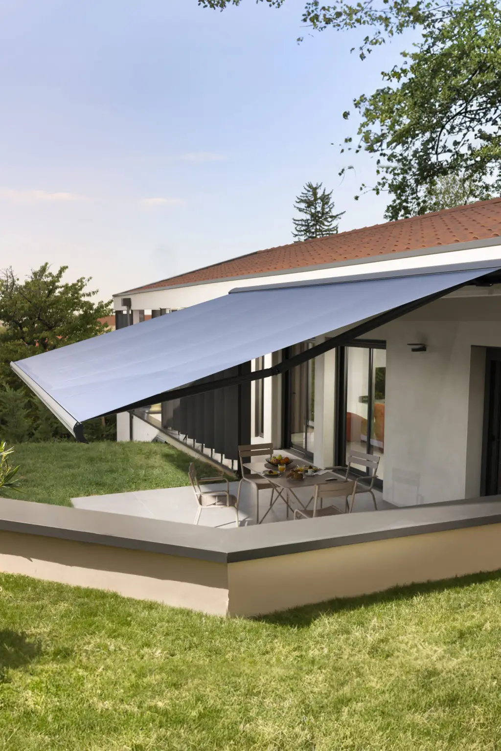 Store banne à toile gris clair déployé au-dessus de la terrasse d’une maison à toit de tuiles ; table et chaises de jardin métalliques, muret, pelouse au premier plan. Format portrait.