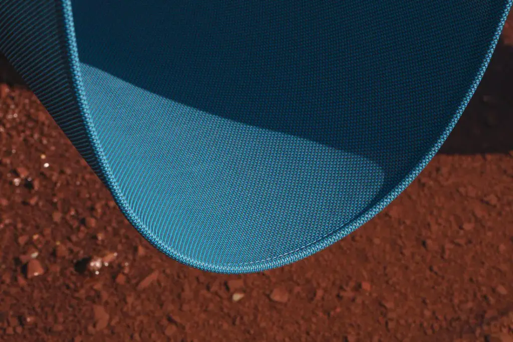 Détail d’une toile ajourée bleu pétrole formant une courbe (assise suspendue), bordée d’un ourlet, au-dessus d’un sol de terre rouge.