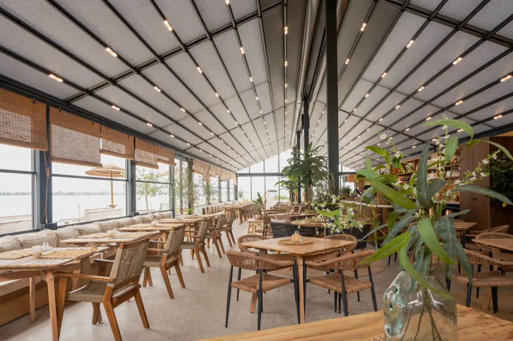 Salle de restaurant de plage sous une grande pergola à structure noire : toiture de toile claire micro-perforée à deux pentes avec rampes de spots, tables en bois, fauteuils en corde, stores en bambou, plantes vertes, mer derrière les vitres.
