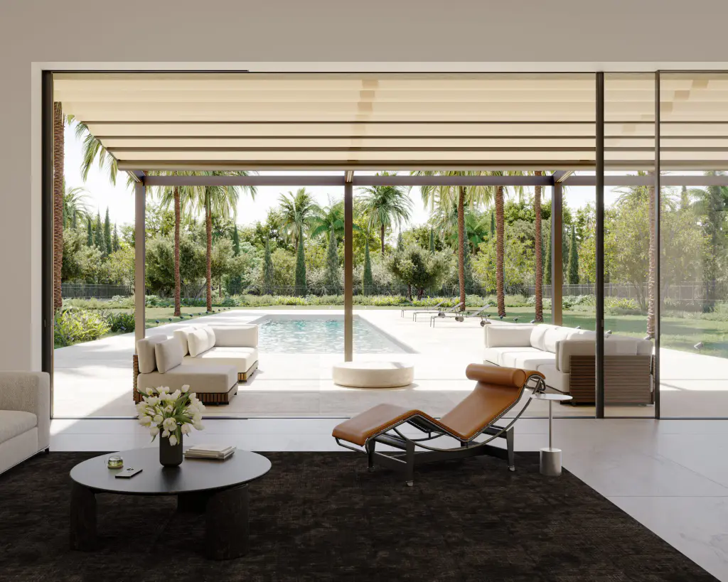 Depuis un salon, vue sur une terrasse couverte d’un vélum de toile claire plissée sous une pergola à fins poteaux bruns ; canapés d’extérieur, piscine, palmiers et cyprès au fond ; chaise longue en cuir et table basse au premier plan.