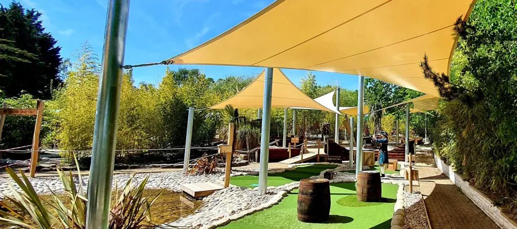 Enfilade de voiles d’ombrage jaune sable tendues sur des mâts en acier galvanisé au-dessus d’un mini-golf paysager : gazon synthétique, galets, tonneaux, bambous, ciel bleu.