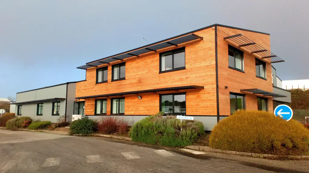 Immeuble de bureaux à deux niveaux en bardage bois clair : un brise-soleil gris anthracite en casquette au-dessus de chaque rangée de fenêtres, massifs d’arbustes, ciel d’orage.