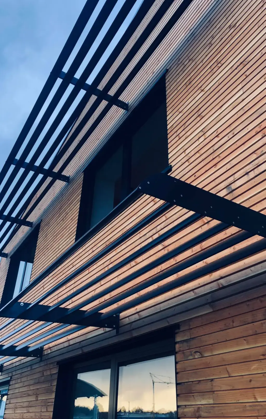 En contre-plongée : deux rangées de brise-soleil en casquette à lames aluminium anthracite fixées entre consoles, sur une façade en bardage bois ; le ciel se reflète dans les fenêtres.